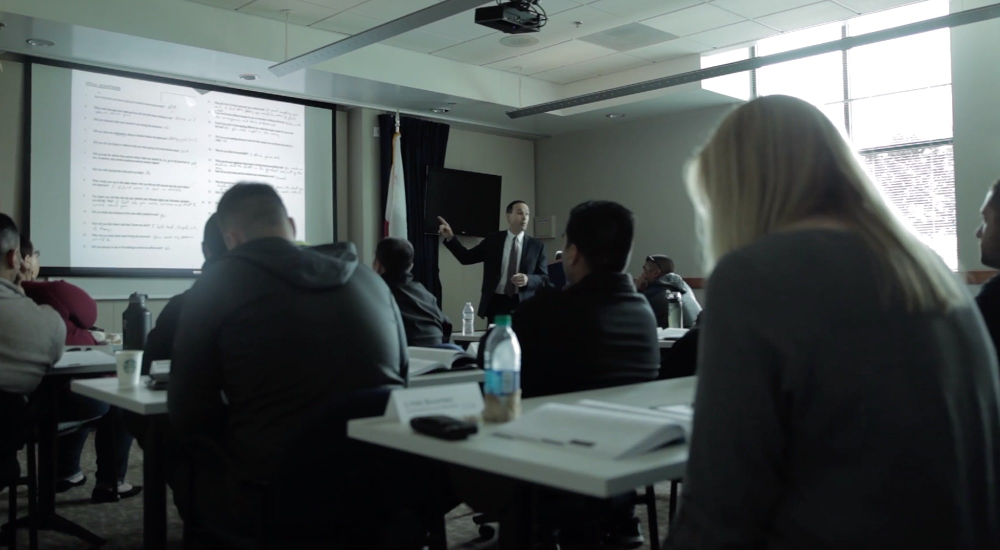 Instructor in suit pointing at projected text while students sit at desks in a classroom.