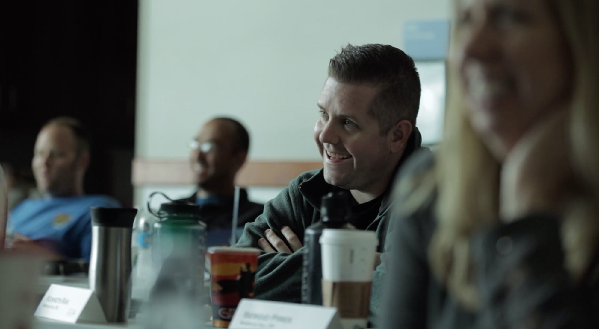 People sitting at a table with drinks, a man in focus smiles and listens in a meeting room.