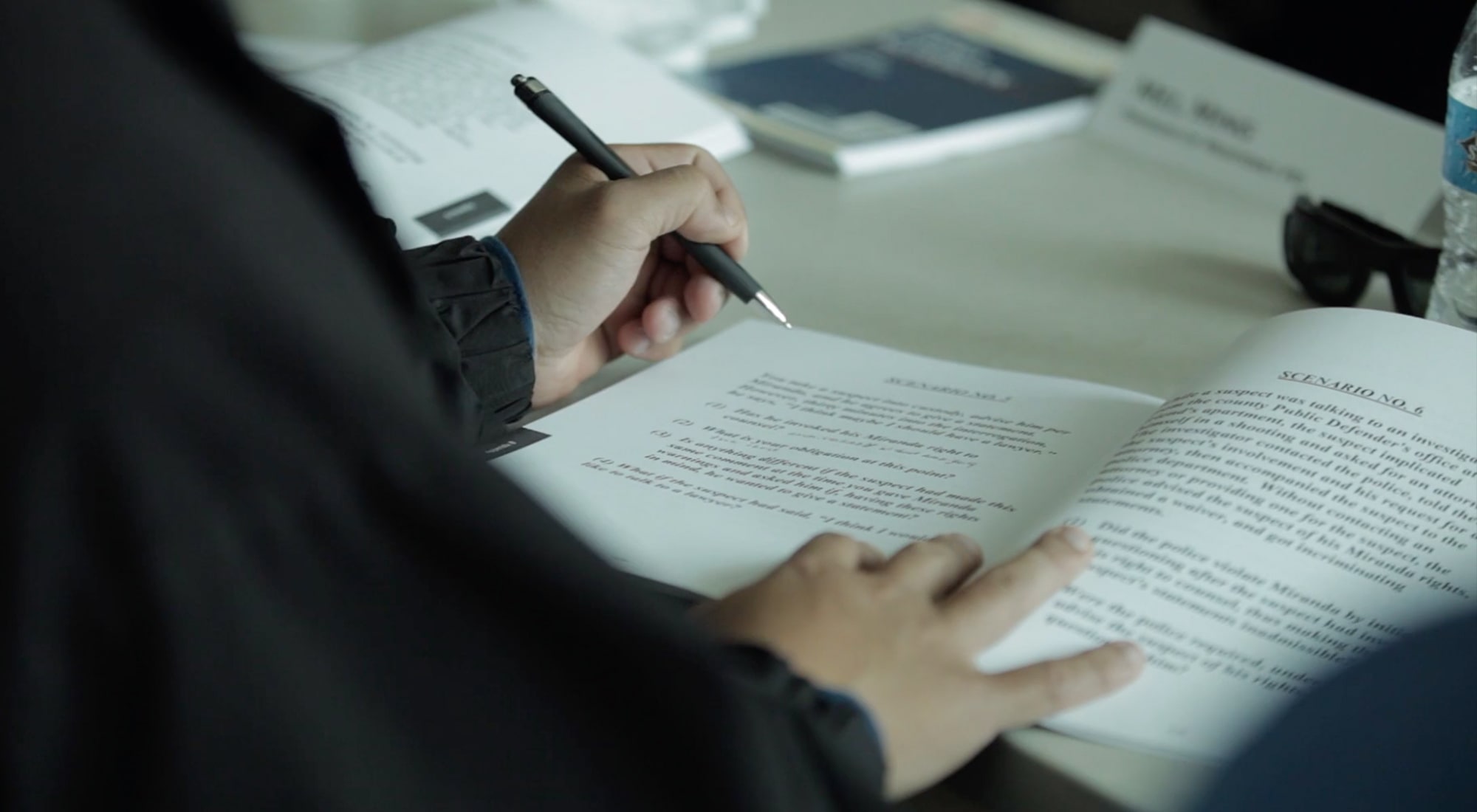 Person holding a pen and reading a booklet with text titled 'Scenario No. 6' at a table.