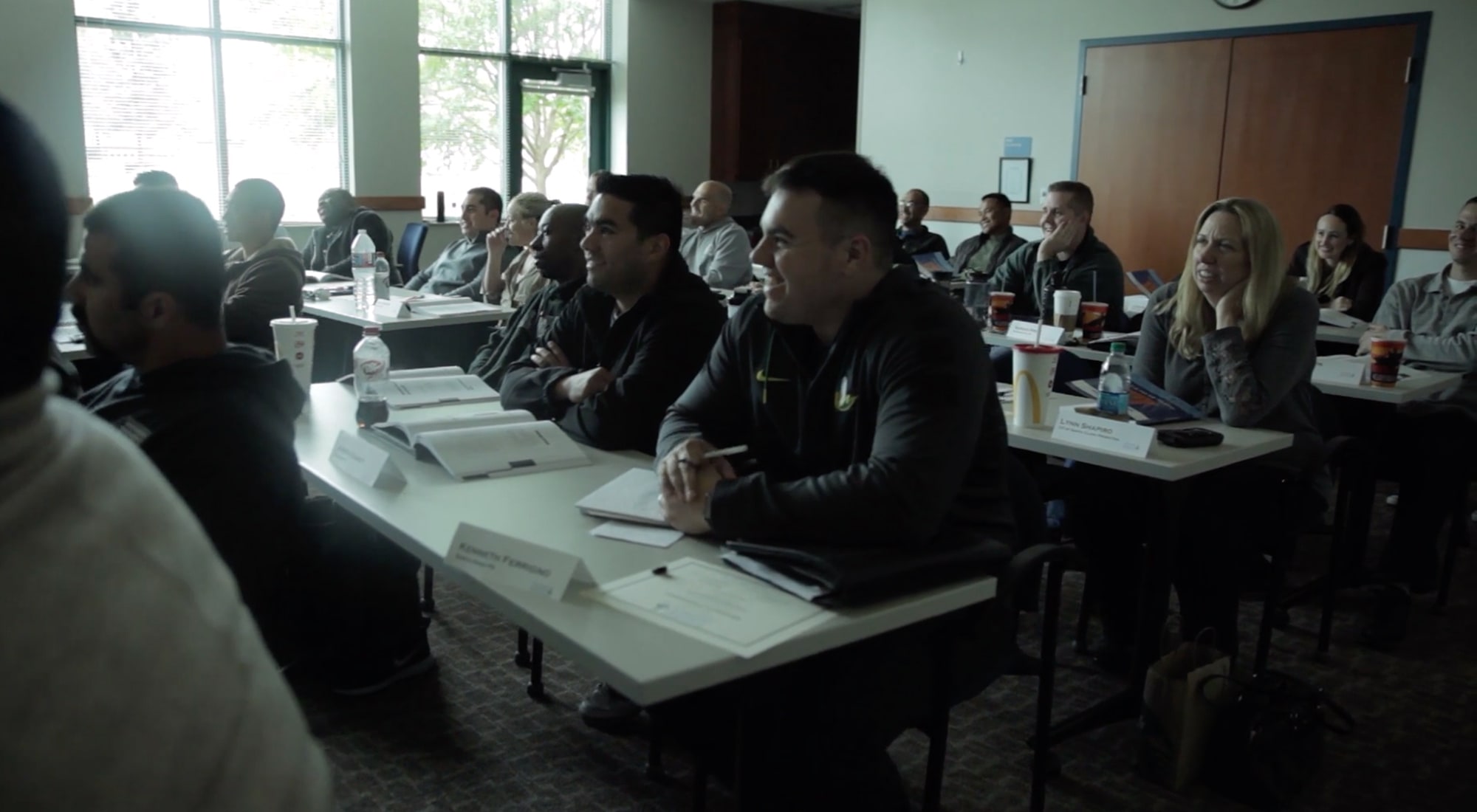Adults seated at desks in a classroom, paying attention and smiling during a session.