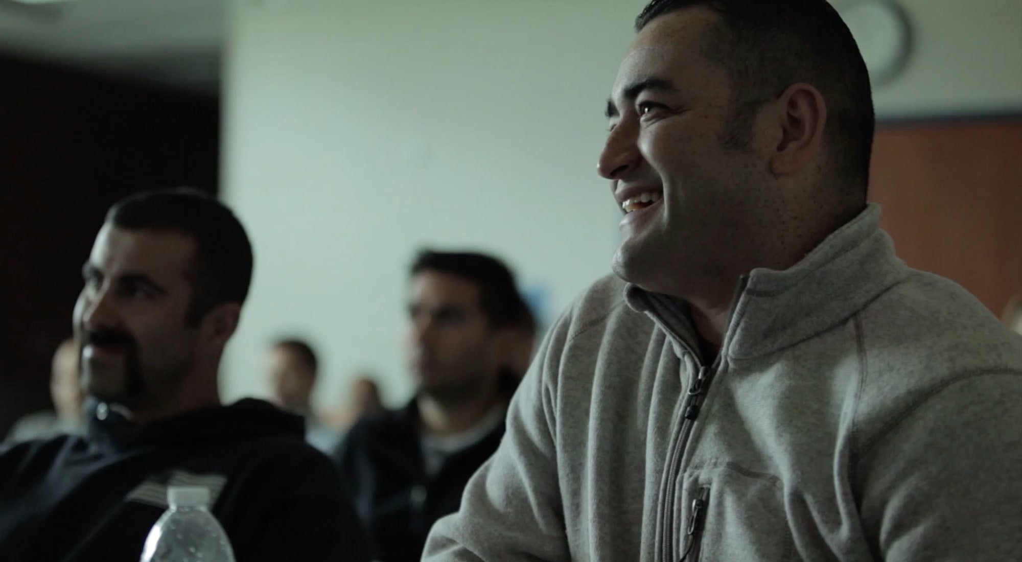 Smiling man in a gray zip-up sweater sits with others, attentively listening in a dim room.