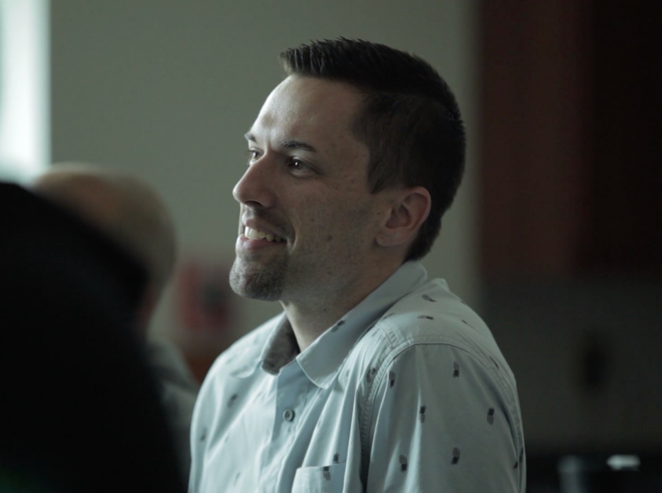 Smiling man with short dark hair and goatee wearing a light shirt with dark patterns indoors.