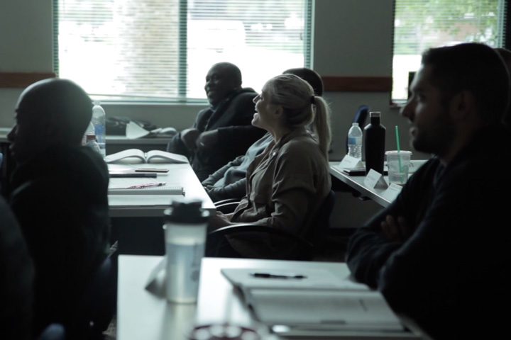 Diverse group of adults seated at desks in a classroom with notebooks and water bottles.