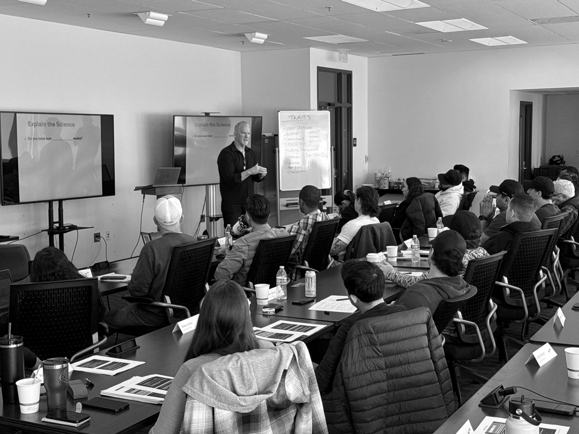 Instructor standing and speaking to a group of people seated in a classroom with presentation screens and notes.