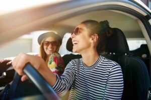 Mulher de blusa listrada e óculos escuros dirigindo um carro, acompanhada de uma amiga de blusa estampada,chapéu e óculos escuros.