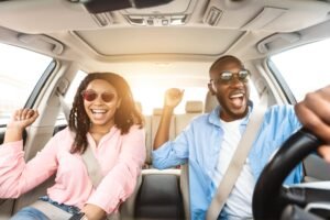 Mulher de camisa rosa e de óculos escuros e um homem de camiza azul e óculos escuros felizes no interior de um carro.