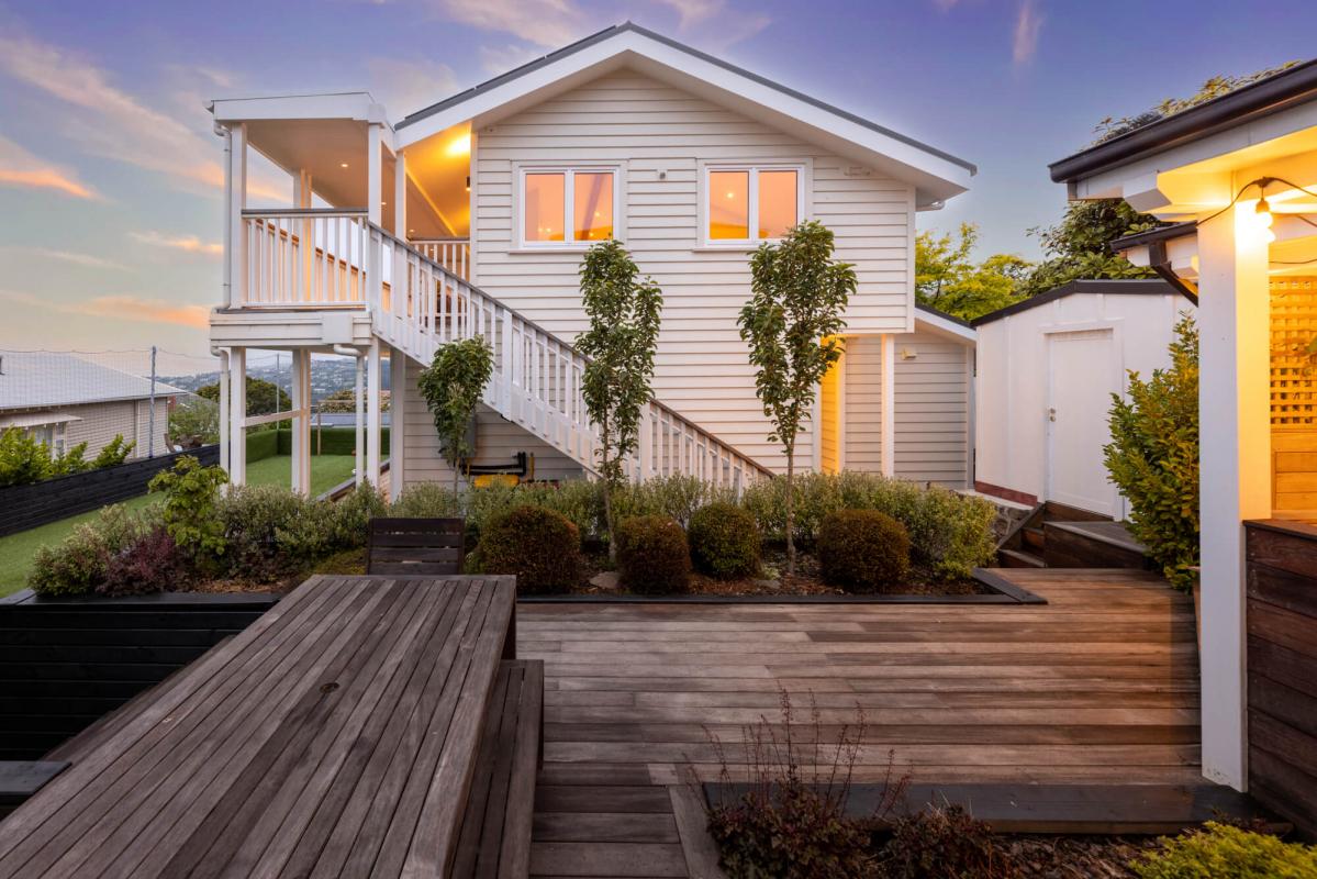 Exterior evening view of the renovated Cashmere Hills character home featuring white weatherboard cladding, wrap-around timber deck, staircase with white balustrades and warm interior lighting at dusk