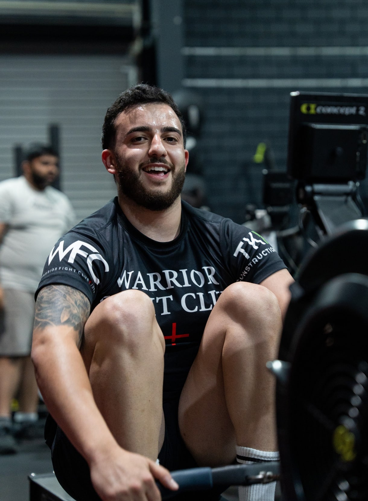 Smiling man in a Warrior Fight Club shirt training on a rowing machine at RevX Fitness Society