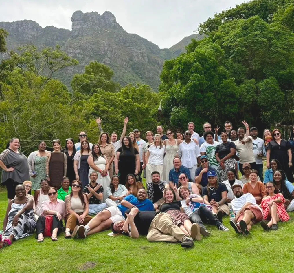 Large diverse group of people posing together outdoors on green grass with trees and mountains in the background.