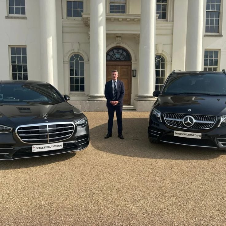 Professional chauffeur standing between two Mercedes executive vehicles outside a formal building
