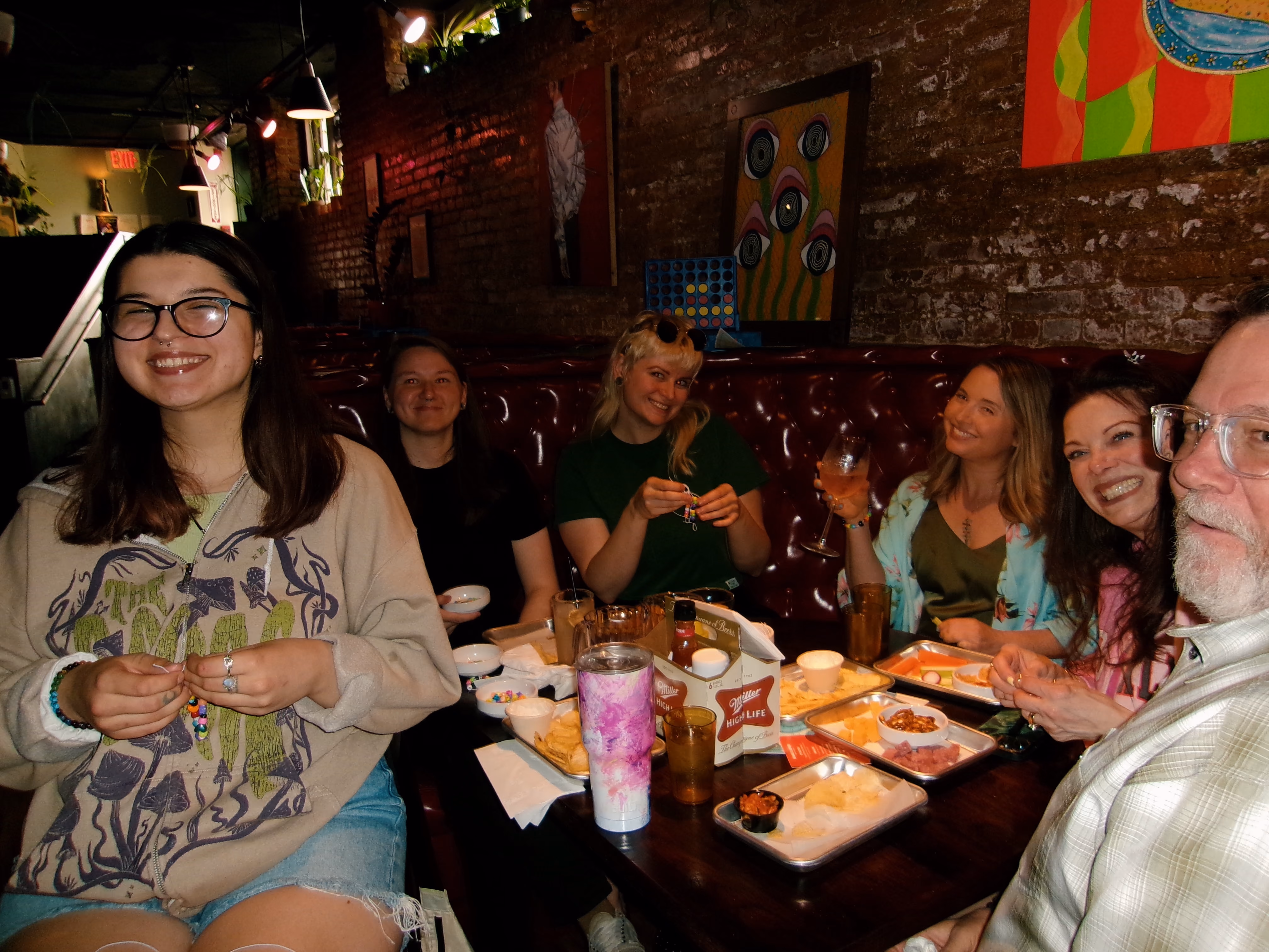 Six people smiling and sitting around a table with snacks and drinks in a cozy, dimly lit restaurant or bar with brick walls and colorful artwork.