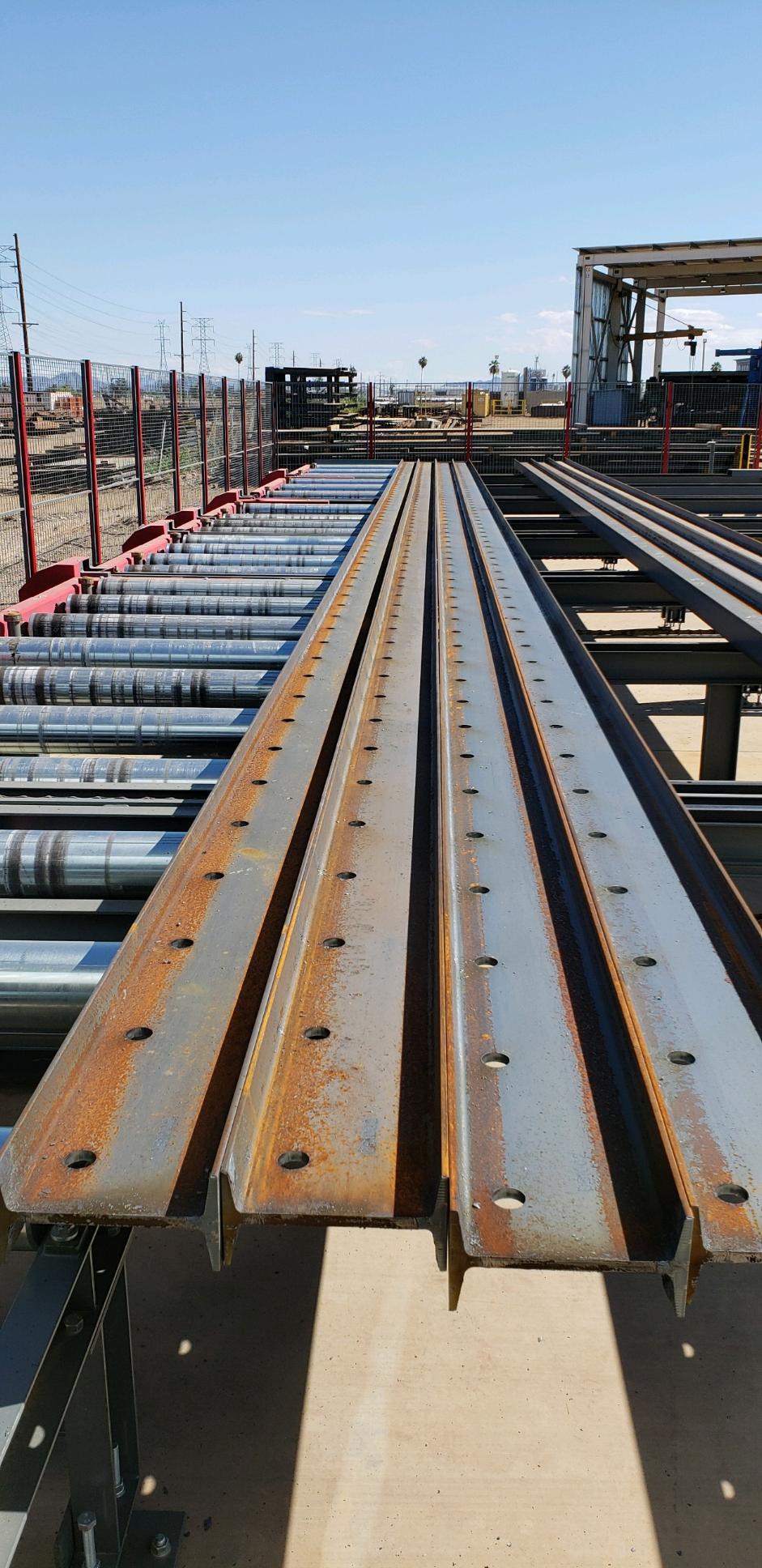 Steel beams with evenly spaced bolt holes moving along roller conveyors in a steel processing facility
