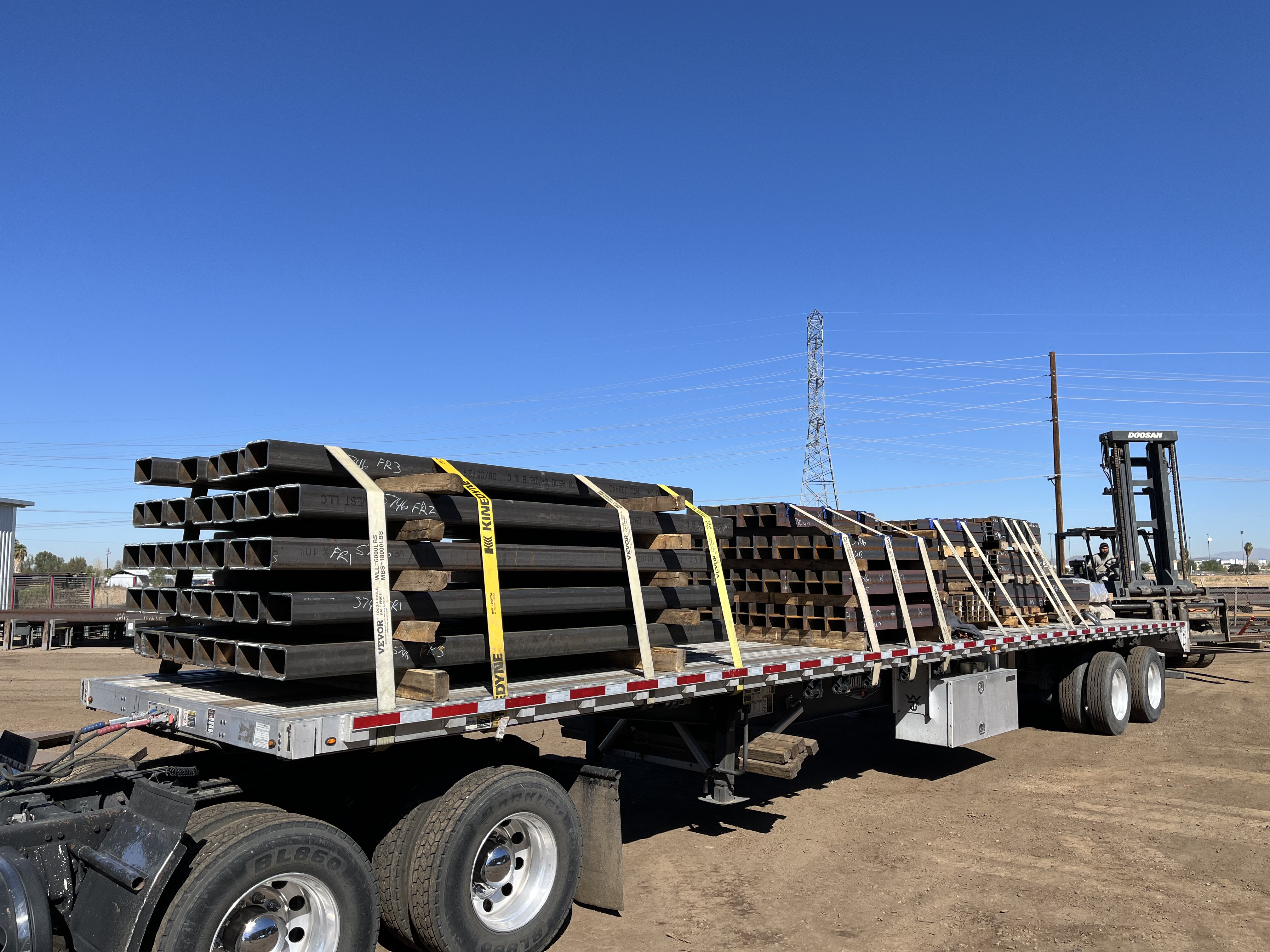 Stacked structural steel sections secured with straps on a flatbed trailer, ready for delivery from a steel processing yard