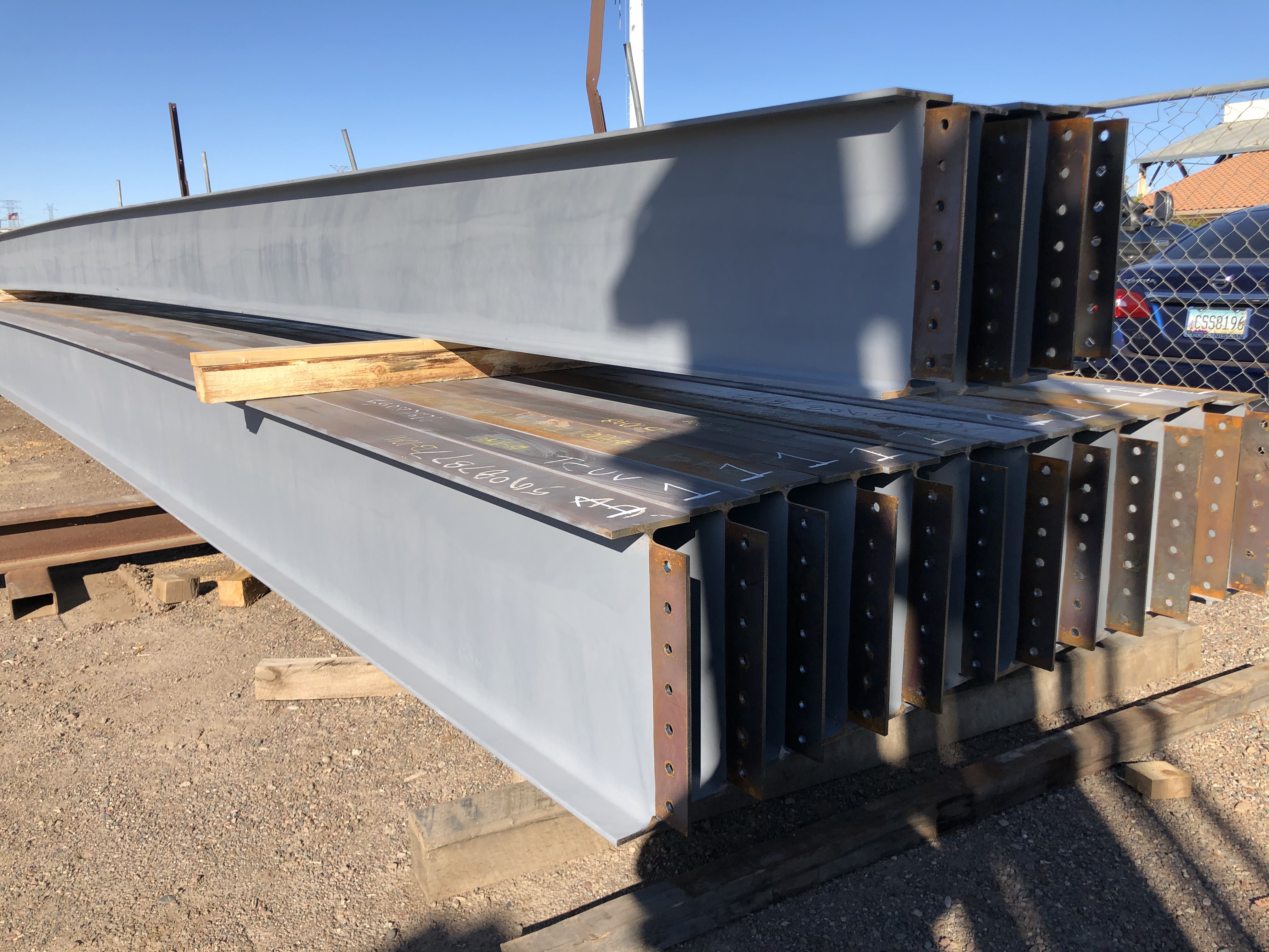 Primed structural steel beams stacked outdoors, showing bolted end plates and protective coating applied for installation