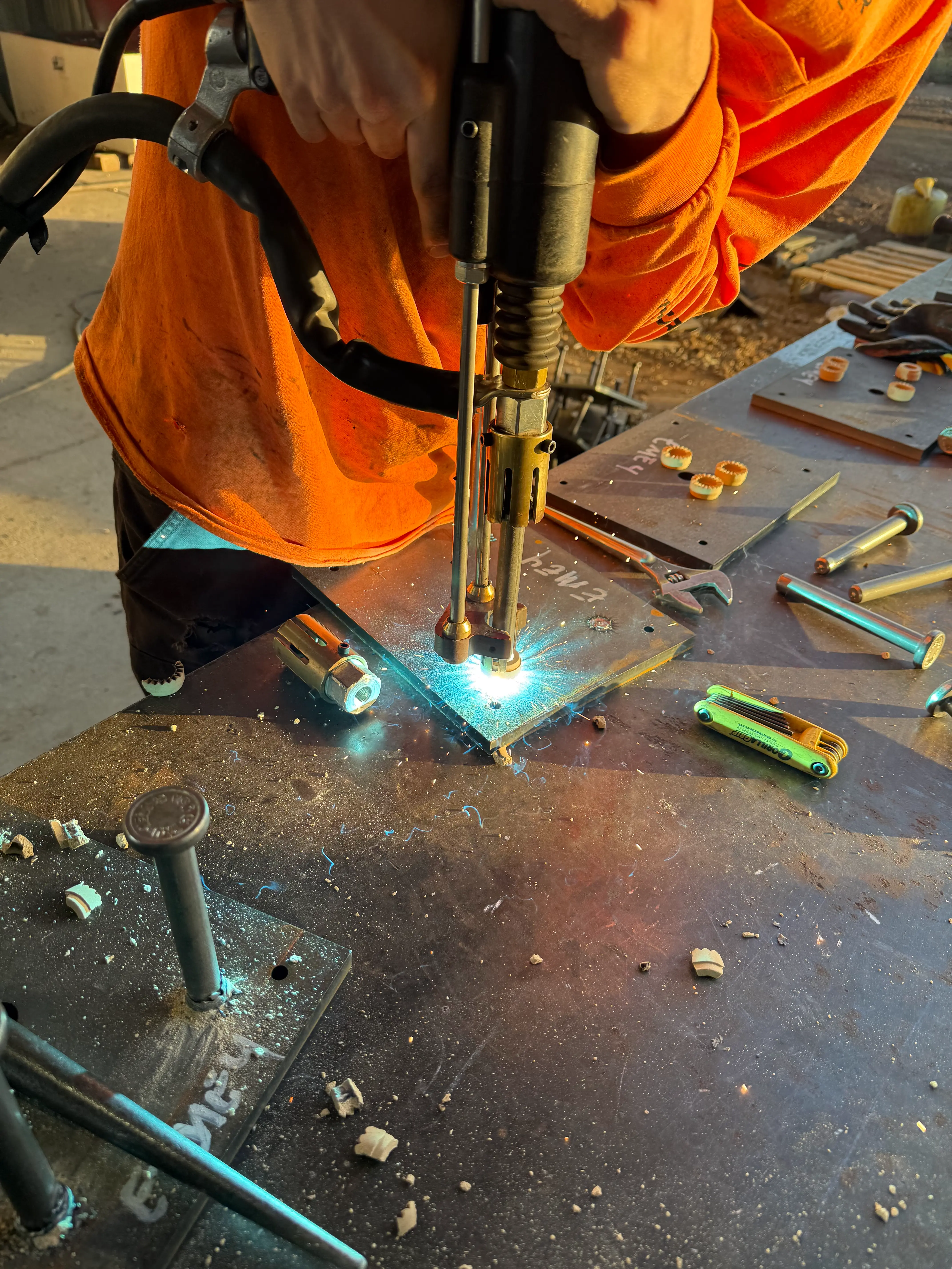 Steel fabrication worker drilling a bolt hole in a steel plate using shop equipment, with metal shavings and tools on the work surface