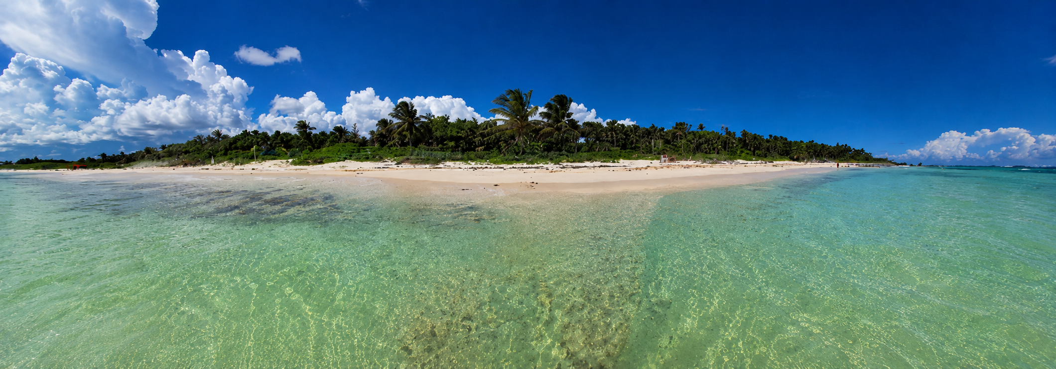 Klares türkisfarbenes Wasser vor einem weißen Sandstrand mit Palmen unter blauem Himmel und Wolken.