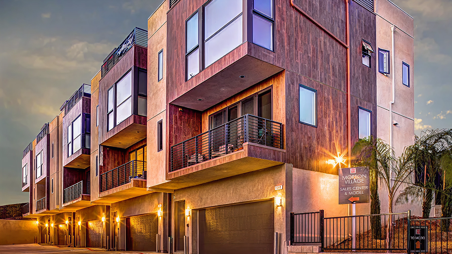 Modern multi-story townhouses at dusk with large windows, balconies, garages, and warm exterior lighting.