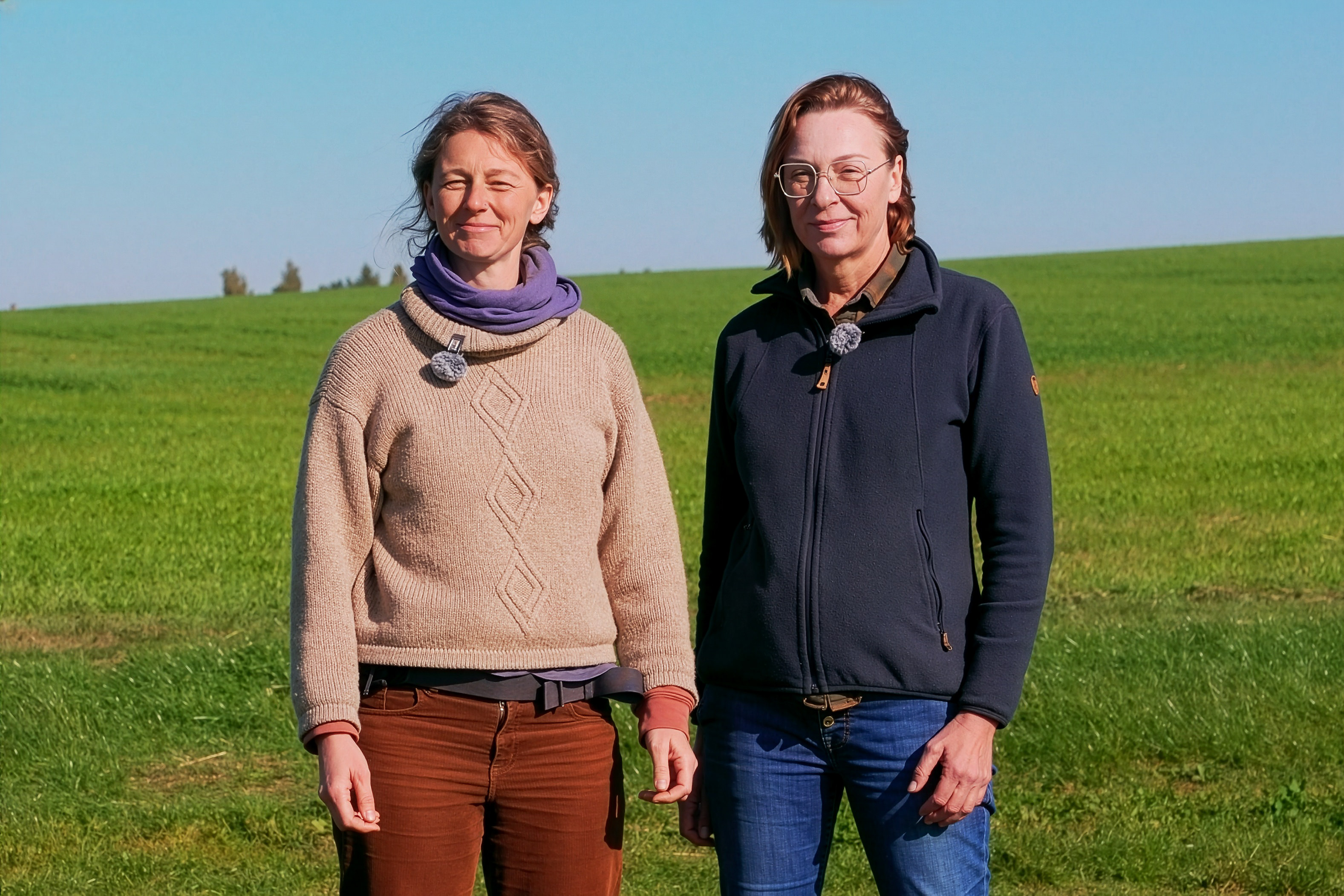 Zwei Frauen stehen auf einer grünen Wiese vor klarem blauem Himmel, tragen Freizeitkleidung und lächeln leicht mit geschlossenen Augen.