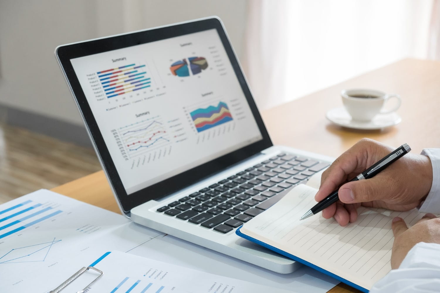 Person holding a pen and writing in a notebook next to a laptop displaying colorful financial charts and graphs, with printed charts on the table and a cup of coffee in the background.