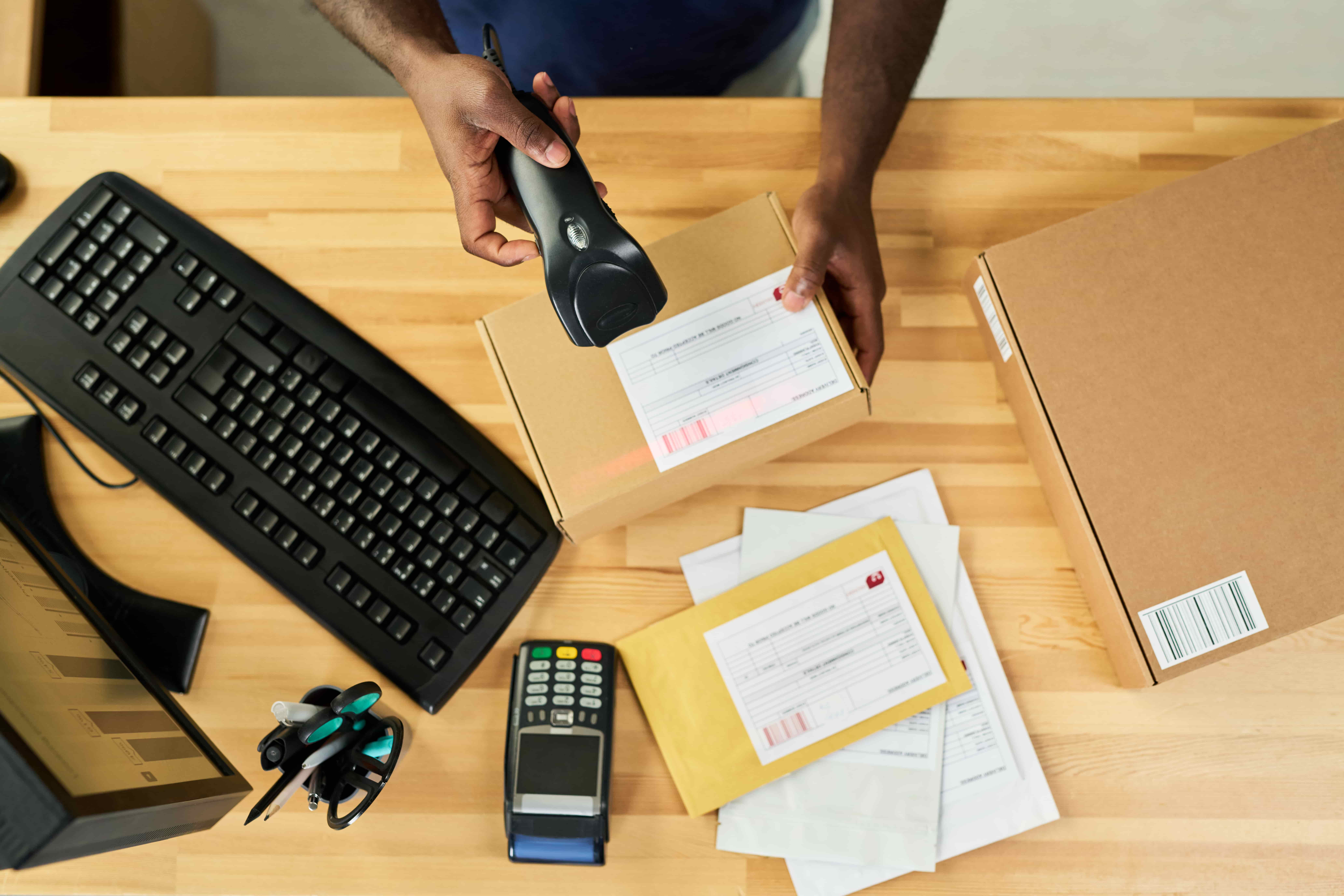 Person scanning a barcode label on a small cardboard box at a desk with a keyboard, monitor, payment terminal, and stacked envelopes.
