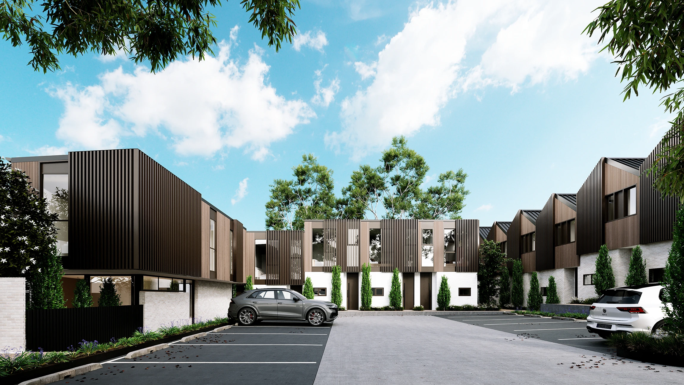 Modern residential townhouse complex with brown paneling, white brick lower walls, green trees, and parked cars under a blue sky with clouds.