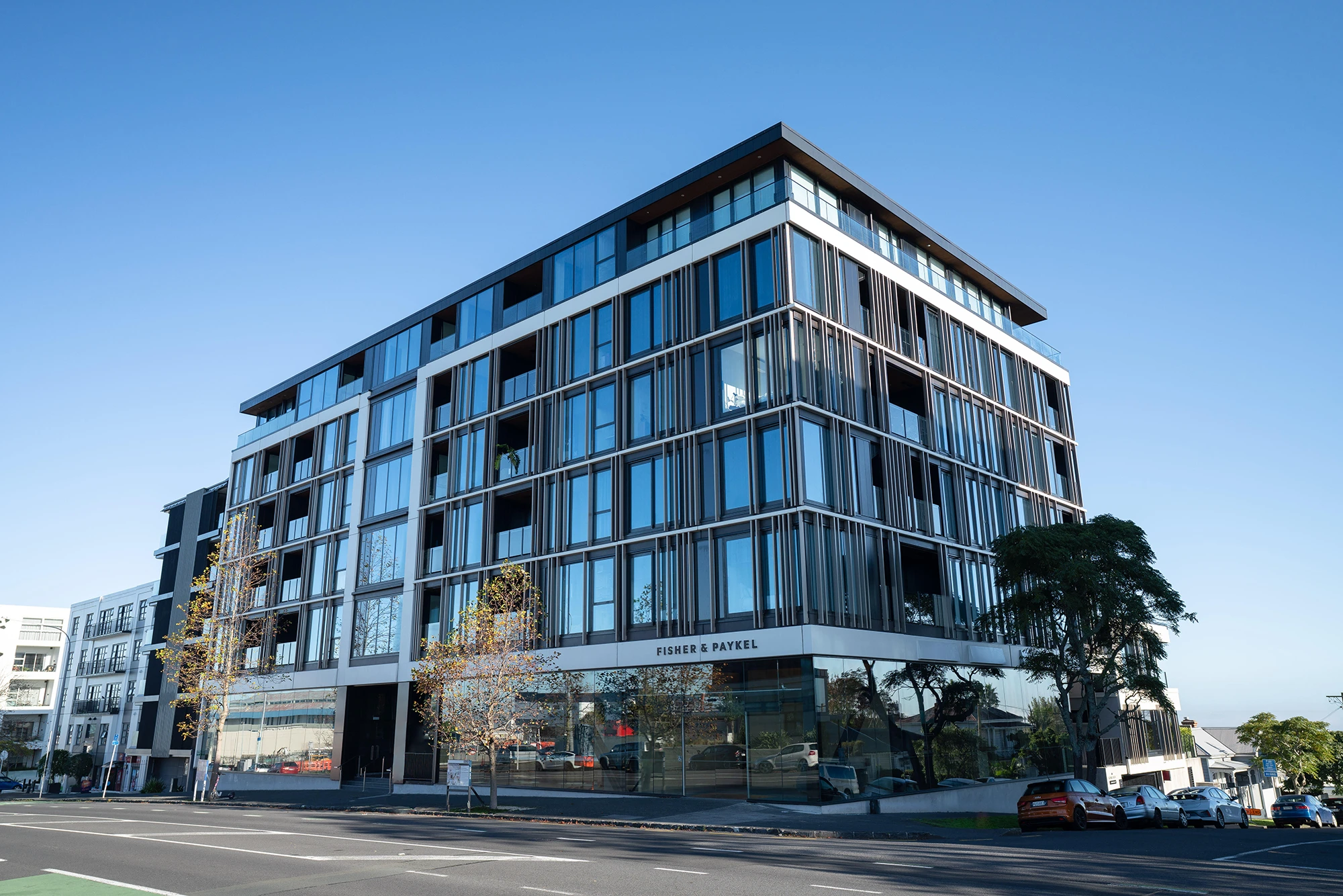 Modern multi-story residential building with large glass windows and balconies on a street corner under a clear blue sky.