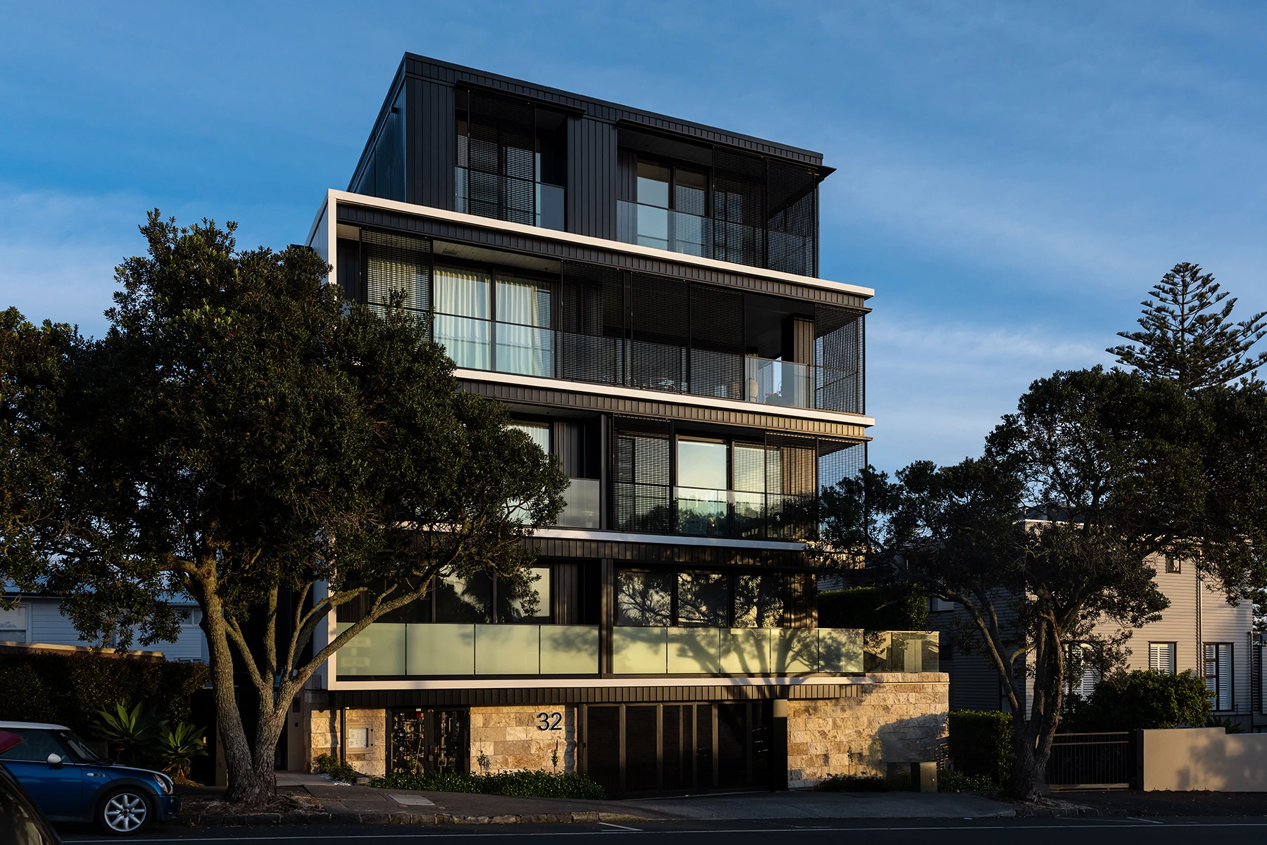 Modern multi-story residential building with dark panels, glass balconies, and stone facade lit by evening sunlight with trees and parked car in front.