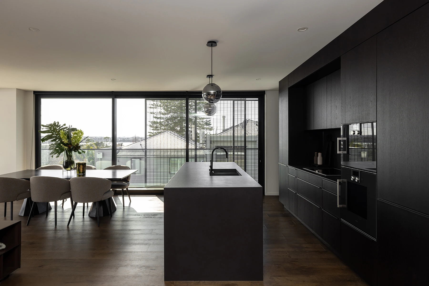 Modern kitchen with a dark island, black cabinetry, a built-in oven, and a dining table with beige chairs by large windows.