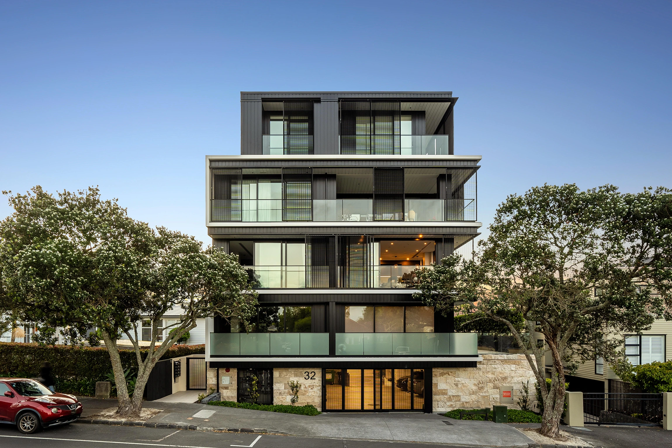 Modern four-story residential building with large glass balconies, black paneling, and stone facade at street level, flanked by trees.