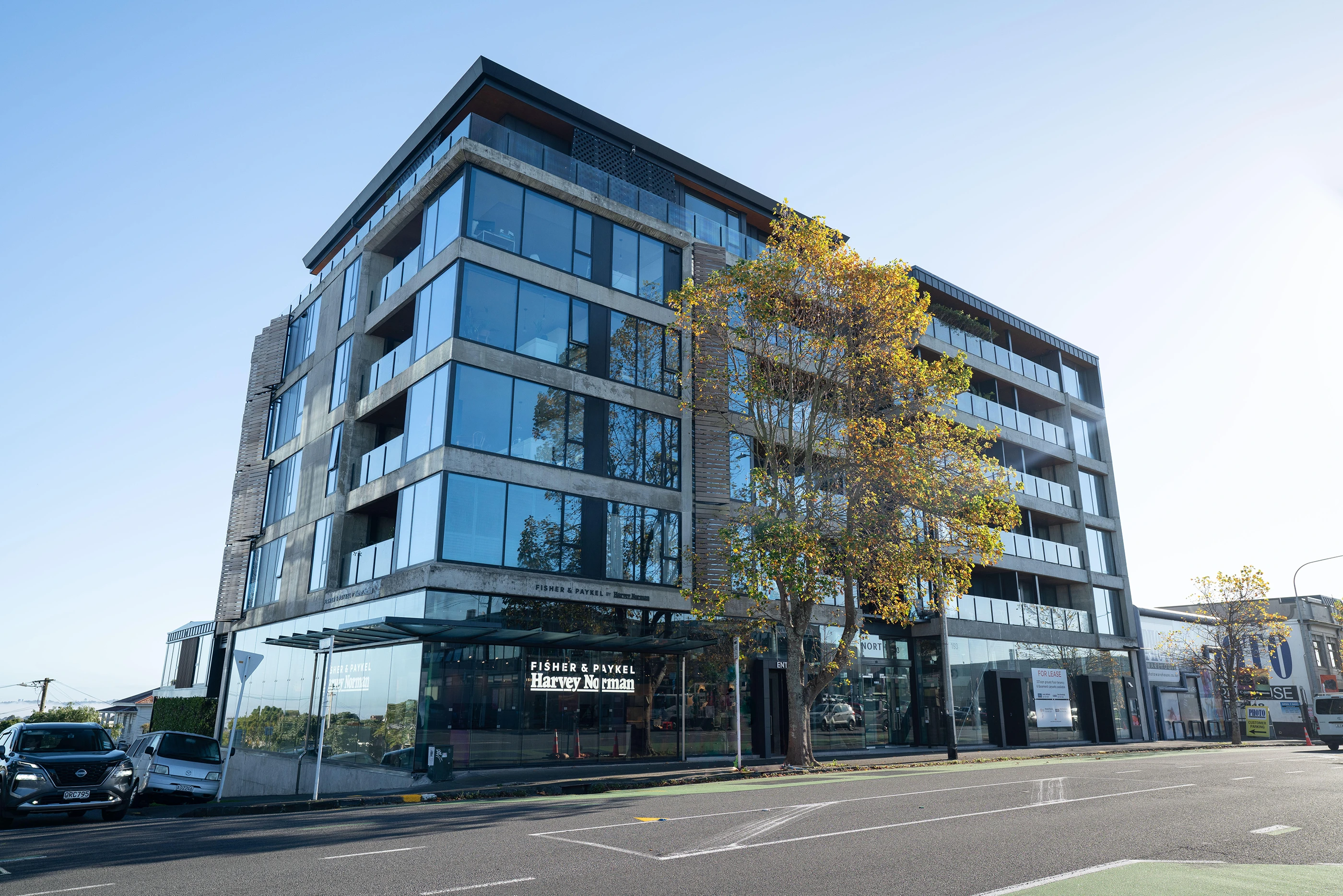 Modern six-story building with large glass windows and balconies, with Fisher & Paykel Harvey Norman store on the ground floor and a tree in front on a sunny day.