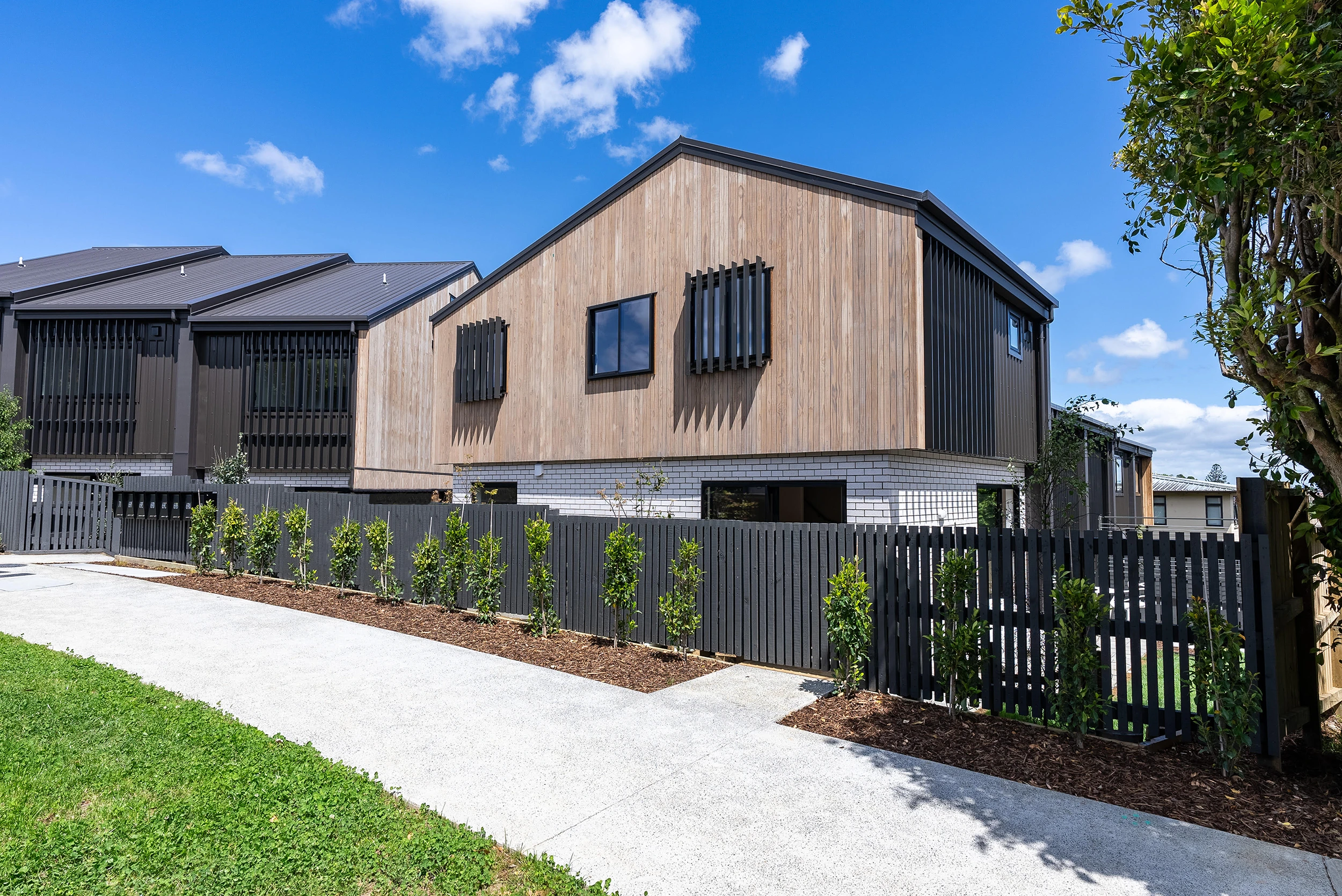 Modern two-story residential building with wood paneling, black accents, and a black fence along a concrete sidewalk.