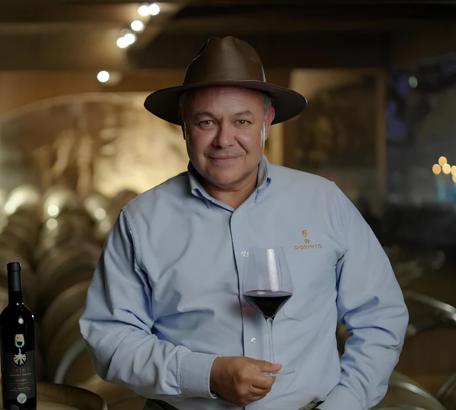 Man wearing a brown hat and light blue shirt holding a glass of red wine in a cellar with wine barrels in the background.