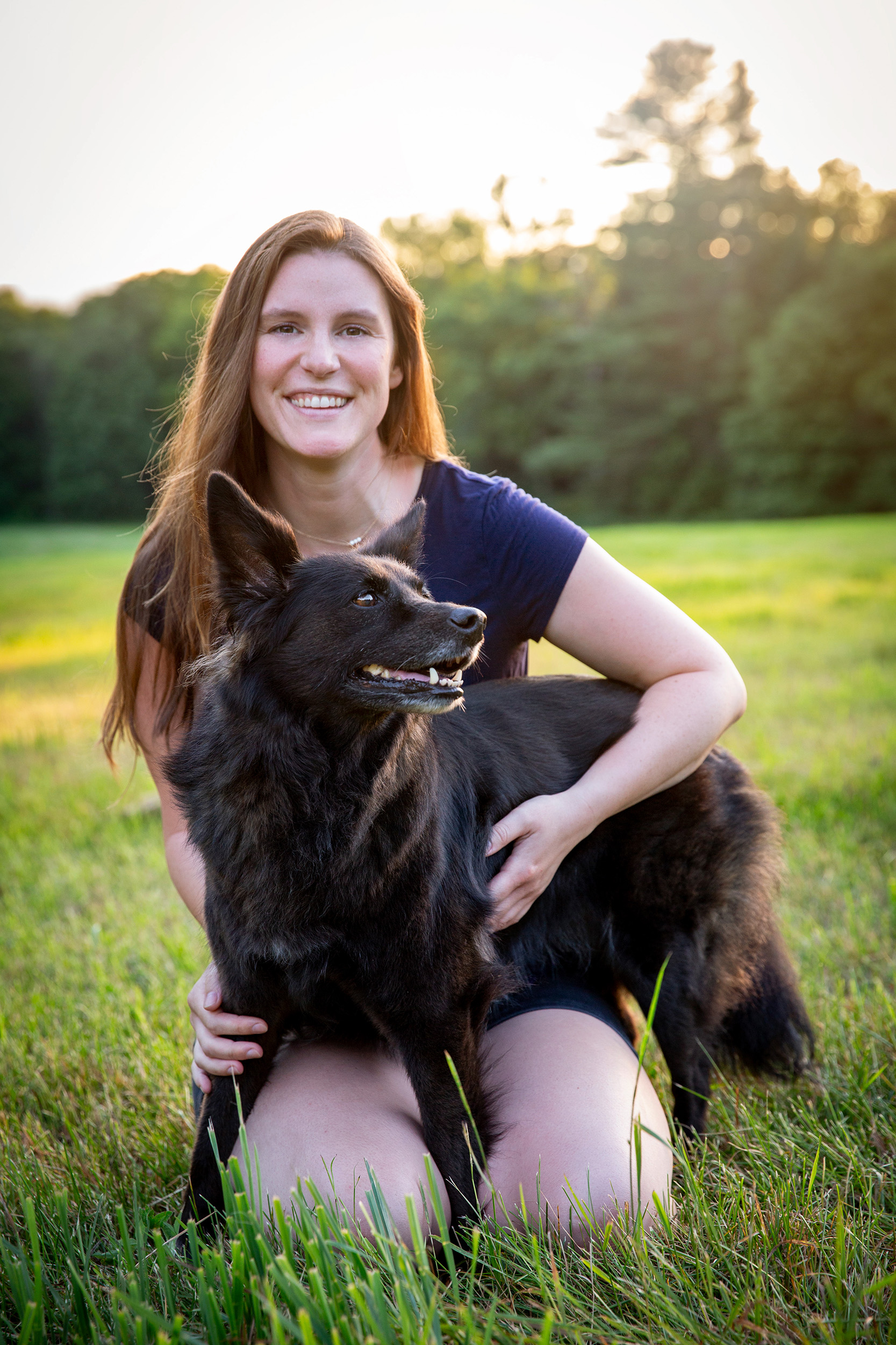 Girl with brown hair kneeling in grass with fluffy black dog across her lap both back lit by sun 