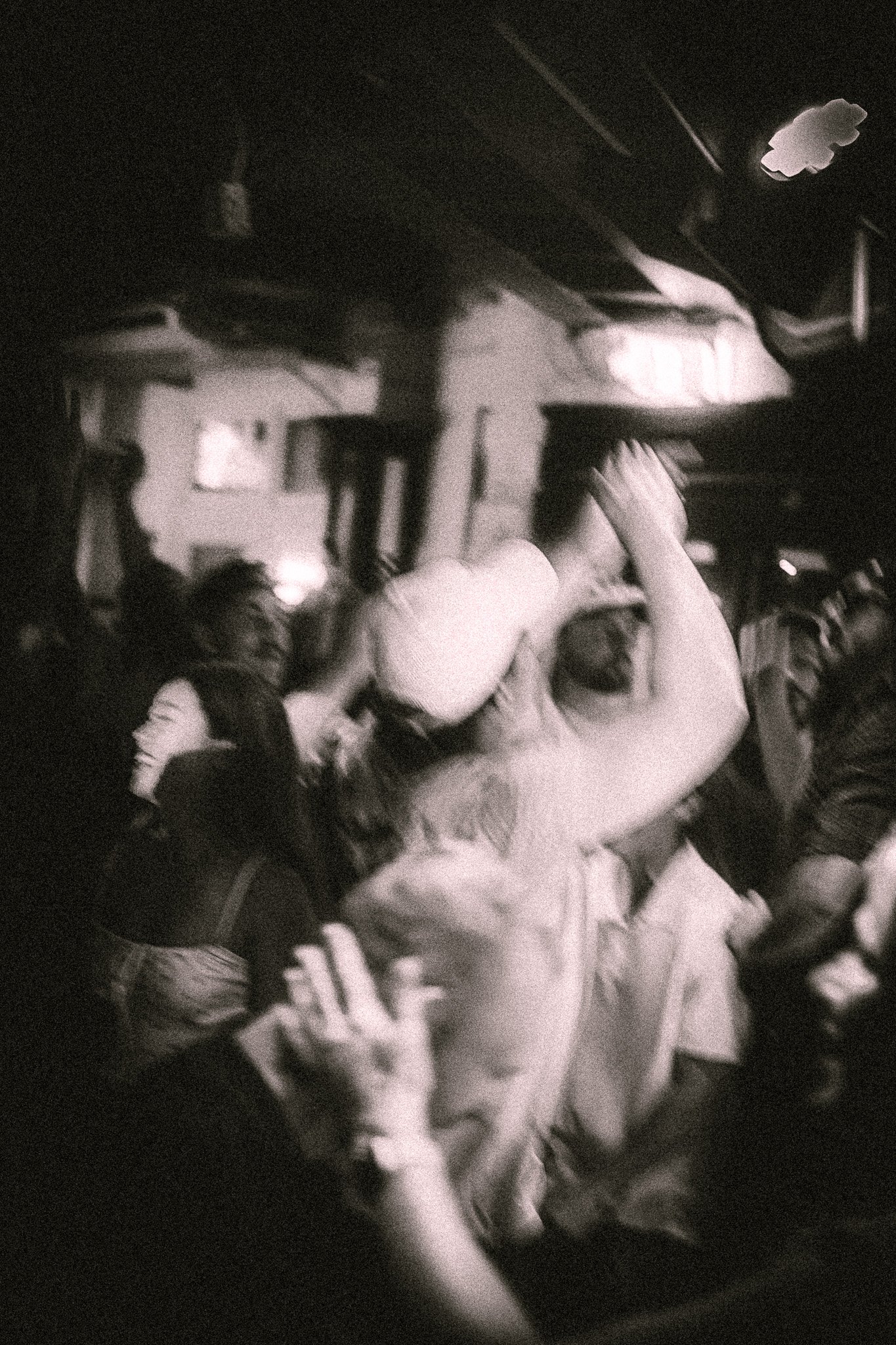 Black-and-white blurred photo of a lively indoor crowd dancing and raising hands listening to REDACTED