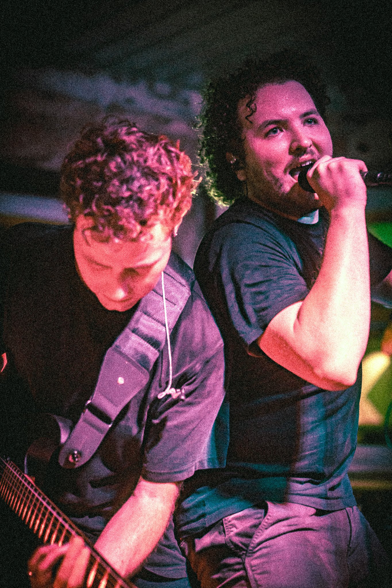 Two musicians from REDACTED performing on stage, one playing guitar and the other singing into a microphone under colorful lighting.