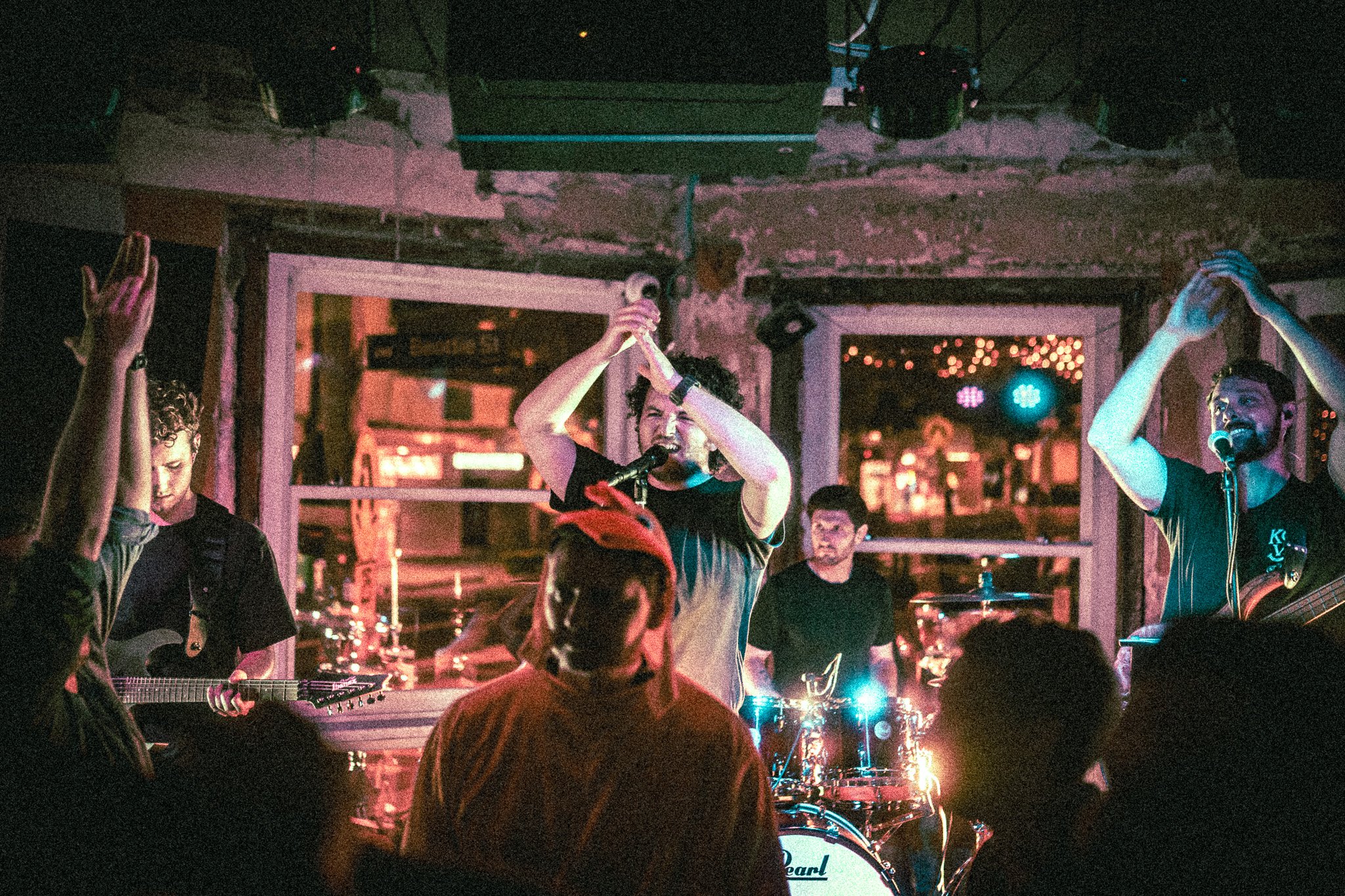 REDACTED Band performing on stage with singer clapping and guitarist, bassist, and drummer playing, audience in foreground.