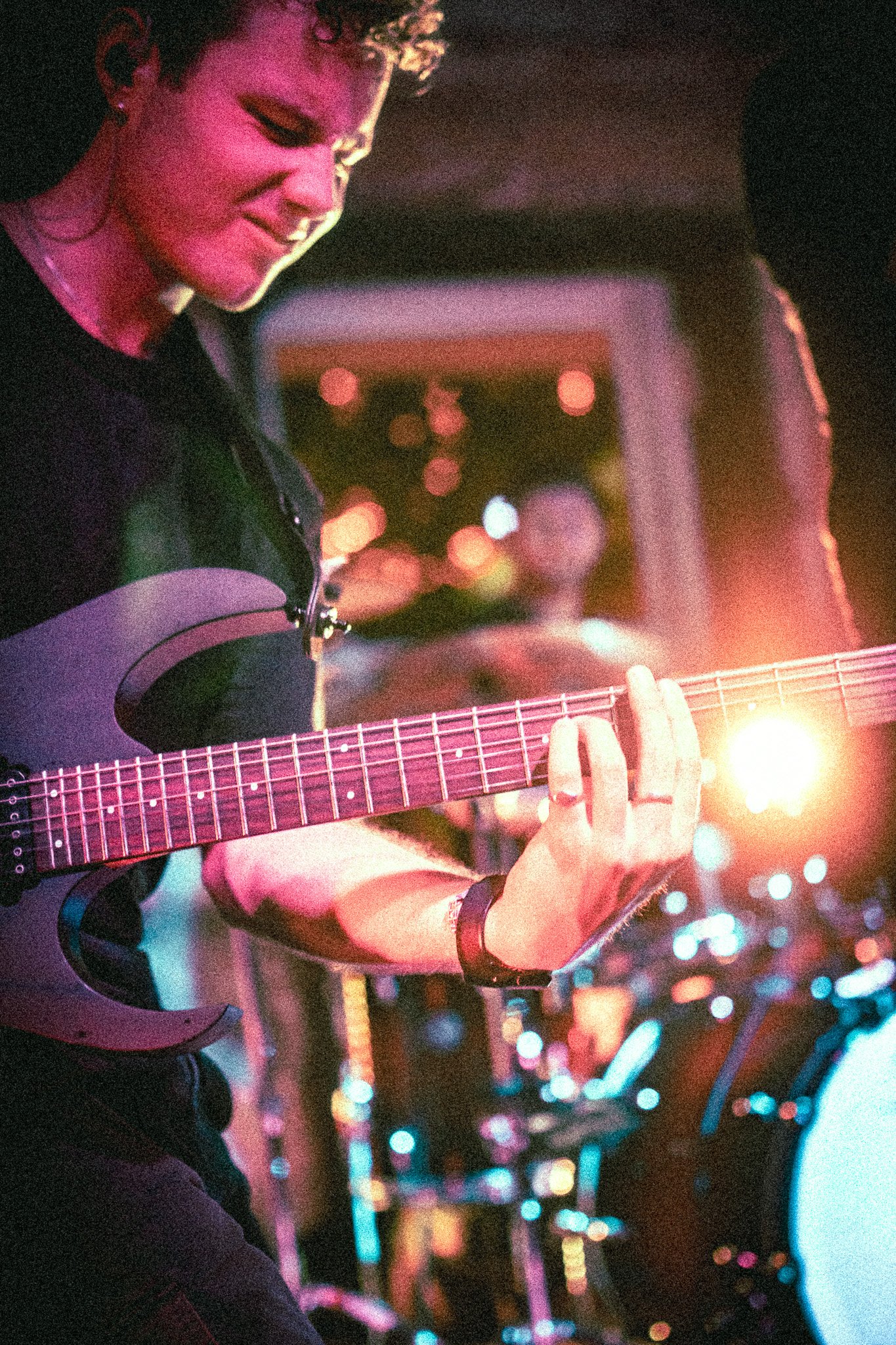 Musician from REDACTED playing electric guitar on stage with colorful stage lights and a drummer blurred in the background.