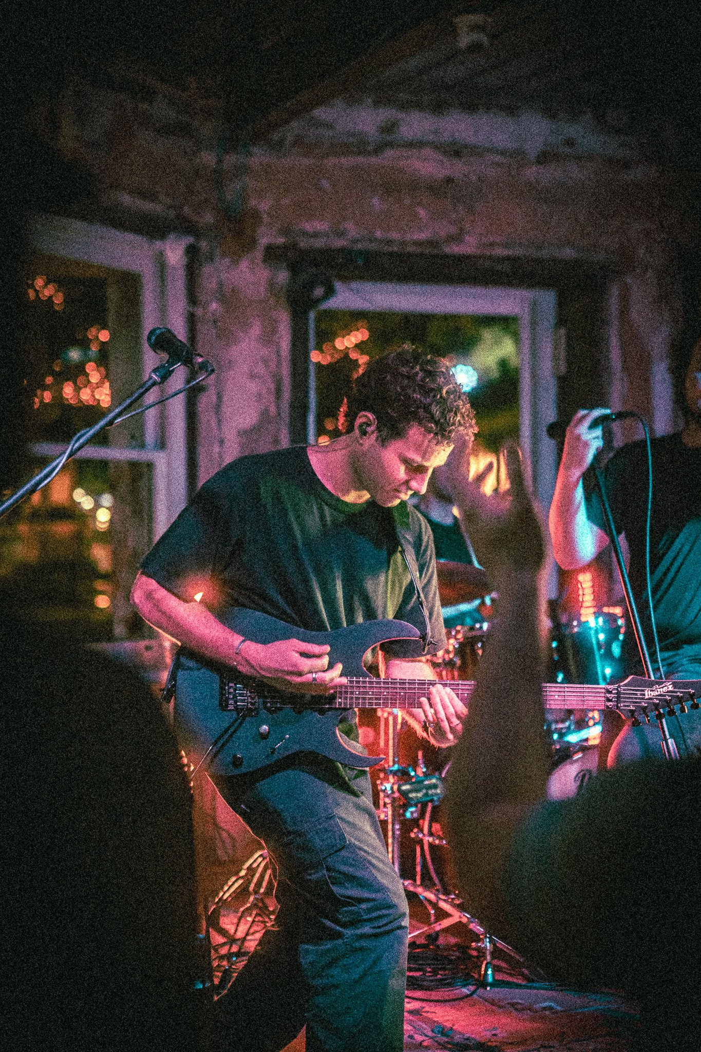 Musician from REDACTED playing electric guitar on stage in a dimly lit venue with raised audience hands.