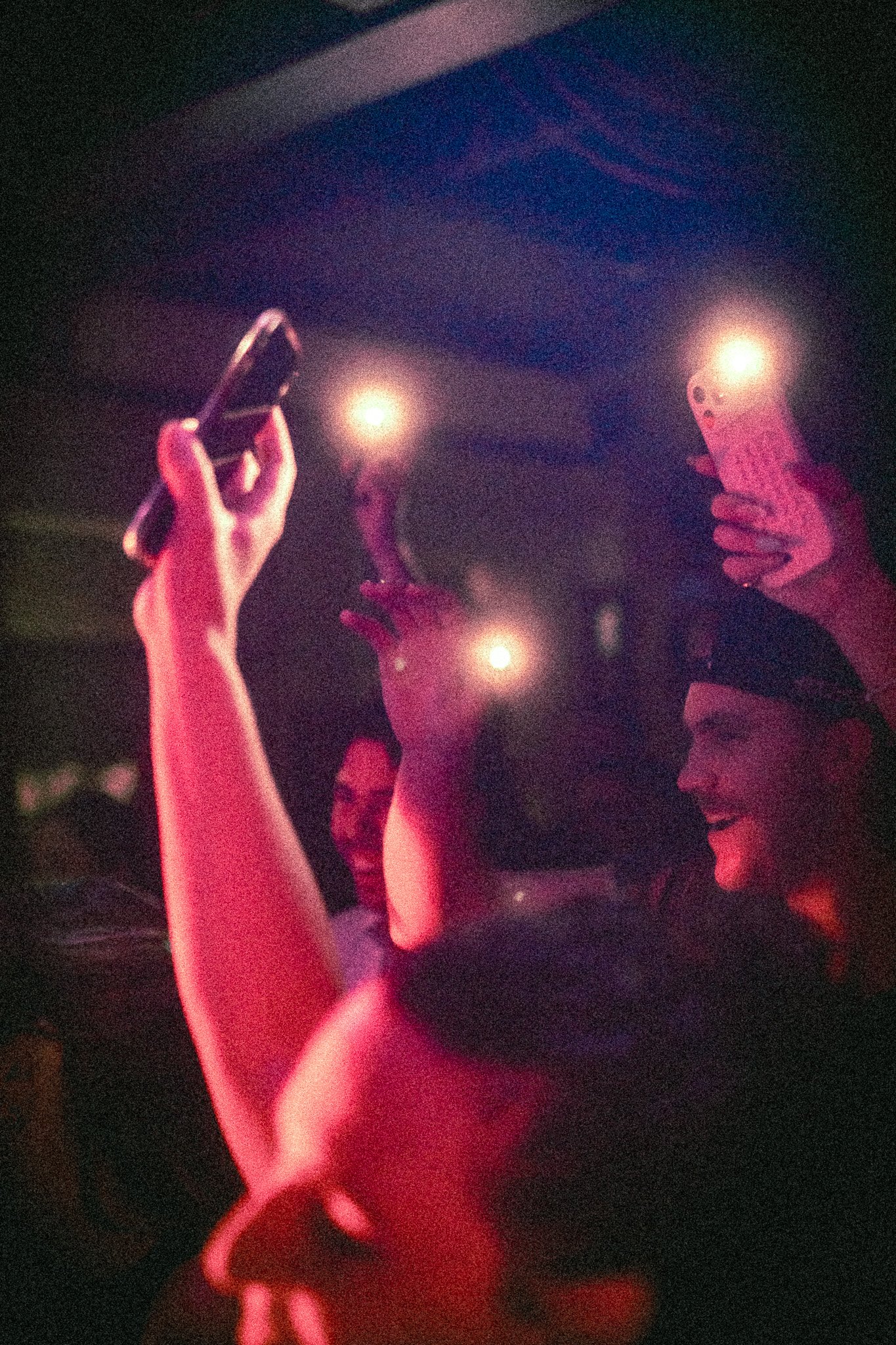 People holding smartphones with flashlights on in a dimly lit crowd, expressing excitement watching REDACTED