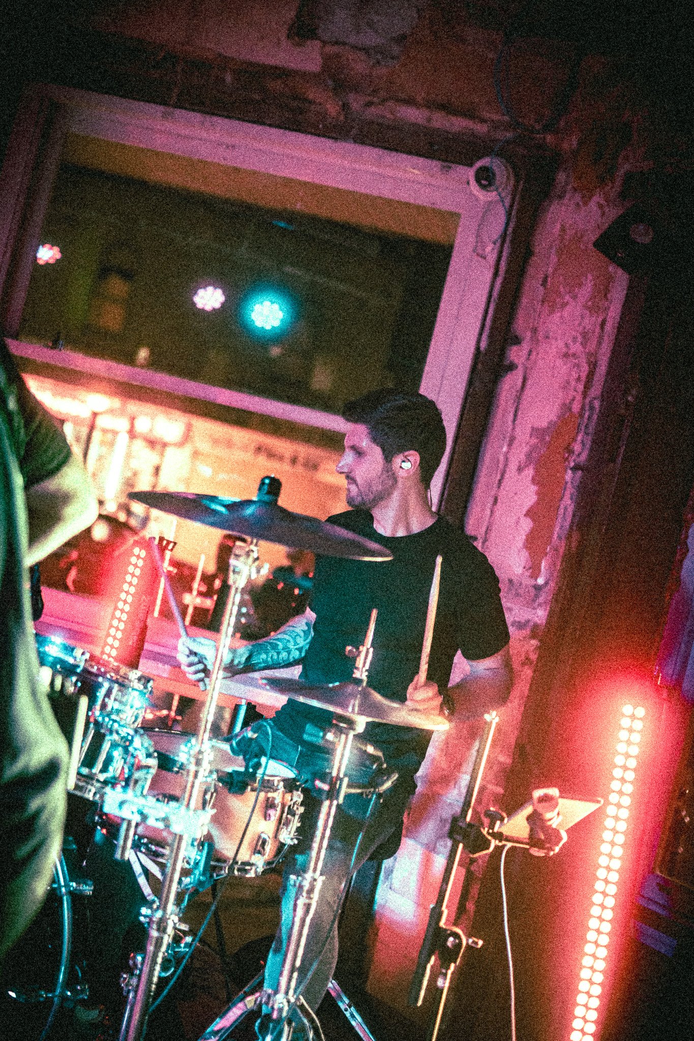 REDACTED musician playing drums in a dimly lit room with colorful stage lights and a window behind him.