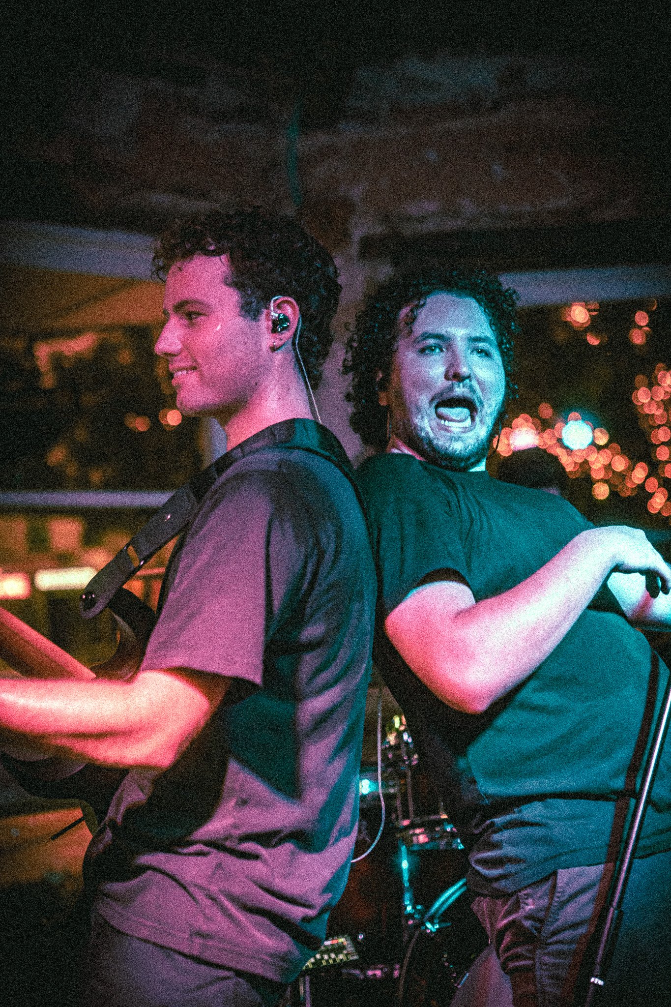 Two male musicians from REDACTED performing on stage, one playing guitar and the other singing into a microphone with colorful stage lights in the background.