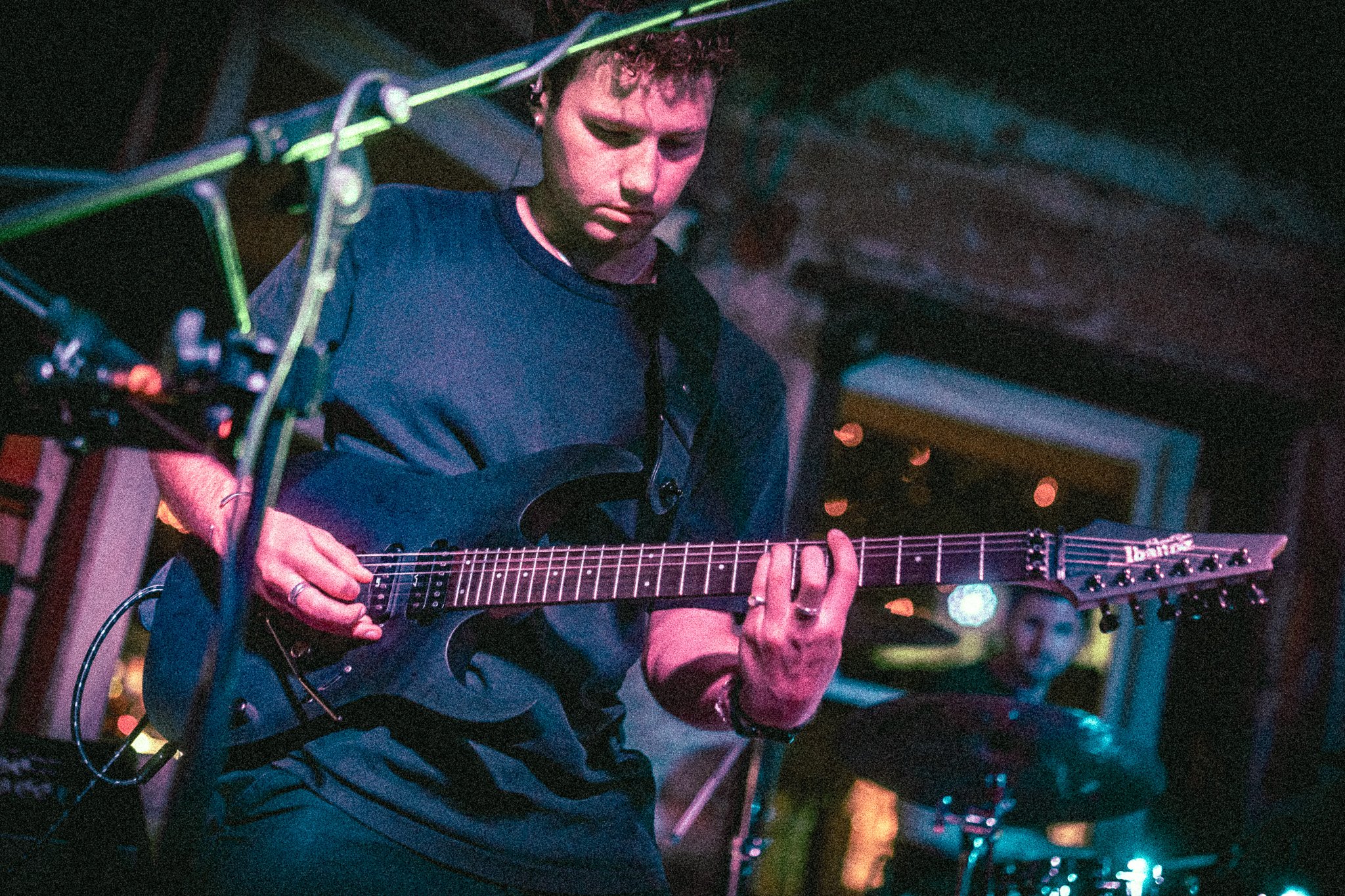 REDACTED playing an electric guitar on stage with a drummer and microphone in the background in a dimly lit venue.