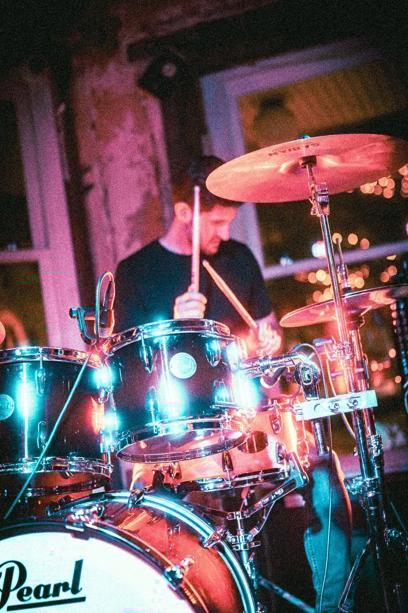 Musician from REDACTED playing a Pearl drum set in a dimly lit room with colorful stage lighting.