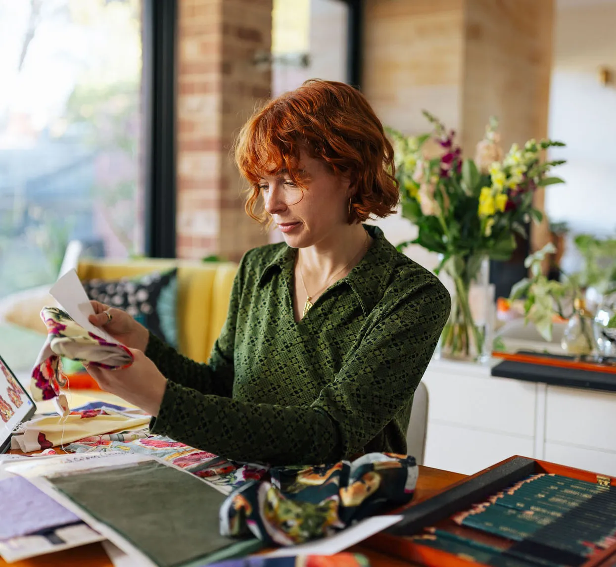 A woman with red hair in a green patterned shirt examines fabric swatches at a table with colorful textile samples and flowers in the background.