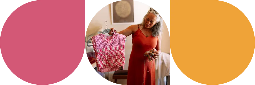 Woman in a red dress holding a knitted pink and white sleeveless sweater on a hanger and yarn in her hands.