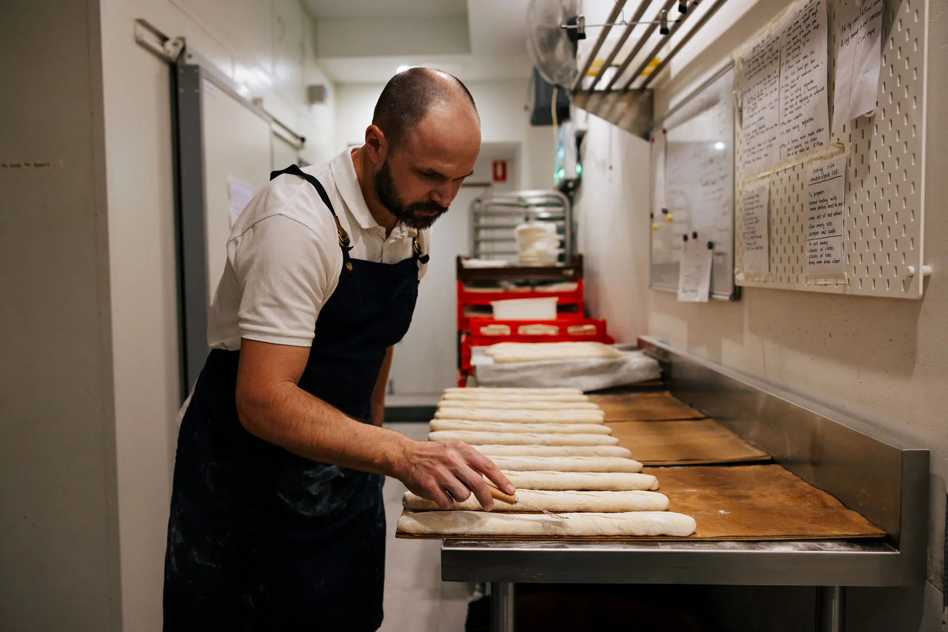 Baker in a white shirt and dark apron scoring long dough pieces on a stainless steel table in a bakery kitchen.
