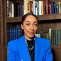 Asha Wadley, Founder of More Than Just Tuition, sits in a blue suit before a bookcase filled with books.