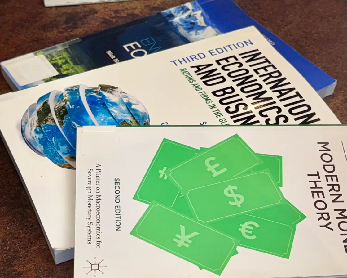 Three economics books on a table beside a green dollar sign, symbolizing financial concepts and market principles.