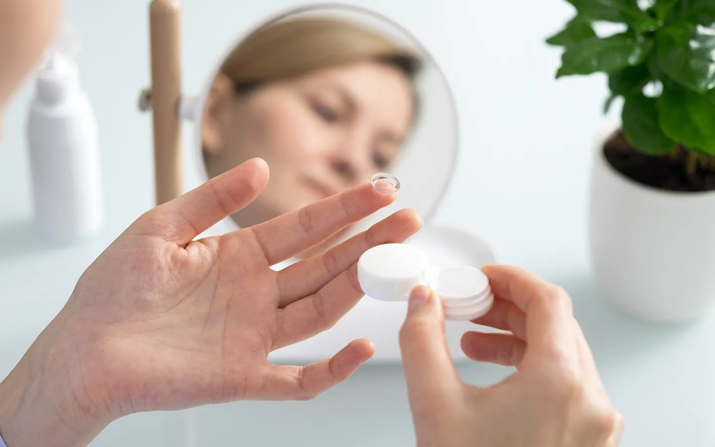 Close-up of a person holding a contact lens on their fingertip and a white contact lens case, with the person's face reflected in a small round mirror.