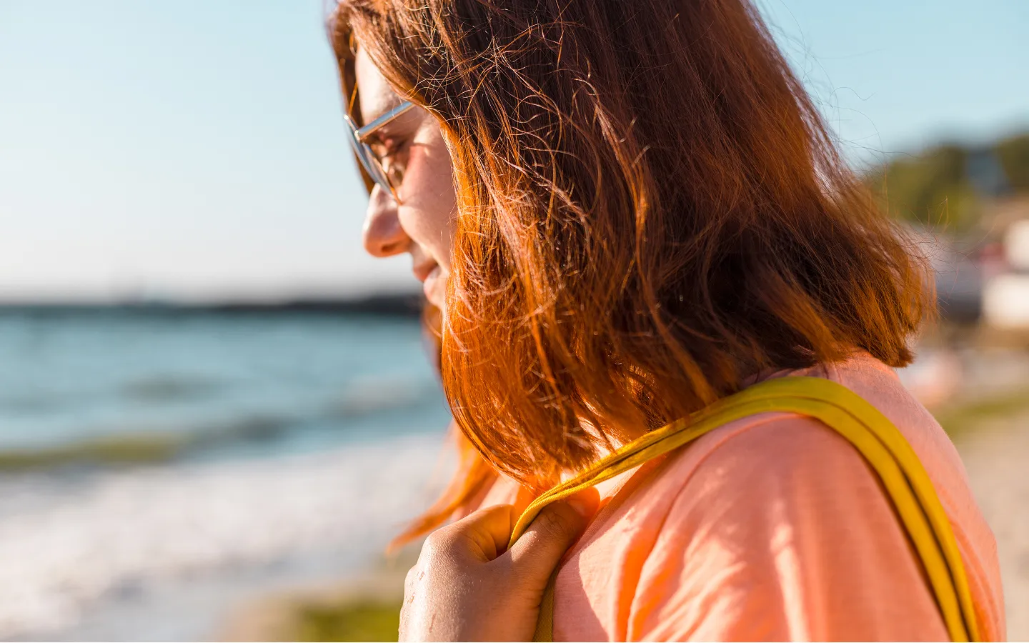 Side profile of a woman with red hair and glasses holding a yellow bag strap at a beach.